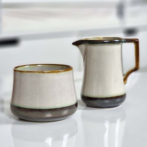 May include: A set of two ceramic kitchenware pieces in a cream color with brown and green accents. The set includes a small pitcher and a sugar bowl.