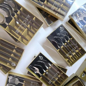 Può includere: Diverse scatole di set di posate vintage. Ogni set include cucchiai, forchette e coltelli con manici marrone scuro e teste argentate. Le scatole sono beige con la scritta "ETON".