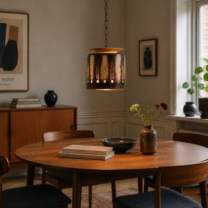 Pode incluir: Uma luminária pendente cilíndrica com um exterior escuro e texturizado, apresentando faixas de luz verticais e uma fileira de recortes circulares na base. A lâmpada pende de uma corrente preta, emitindo um brilho quente sobre uma mesa.