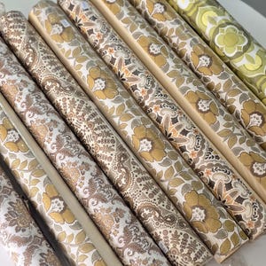 May include: A collection of eight rolls of wallpaper arranged side by side. The rolls feature various patterns in shades of mustard yellow, brown, cream, and olive green, including floral, paisley, and geometric motifs printed on paper surfaces.