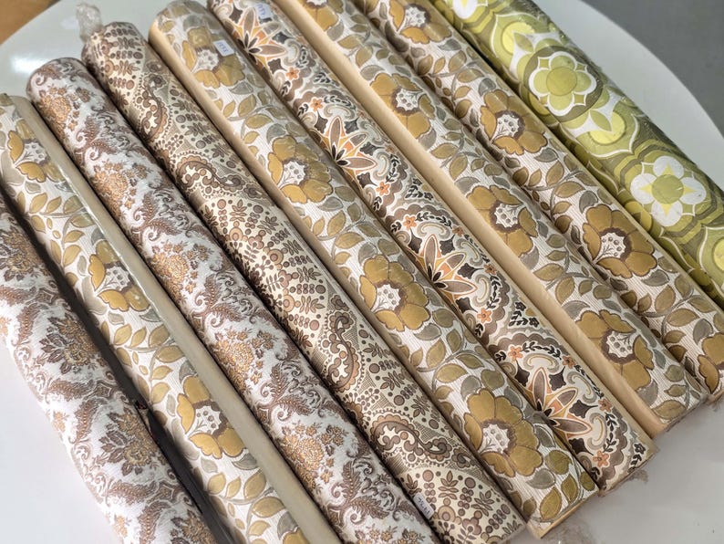 May include: A collection of eight rolls of wallpaper arranged side by side. The rolls feature various patterns in shades of mustard yellow, brown, cream, and olive green, including floral, paisley, and geometric motifs printed on paper surfaces.
