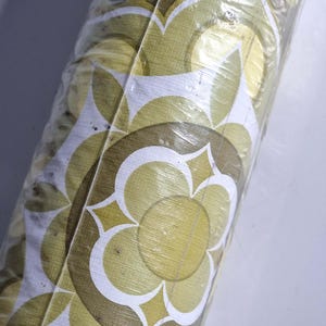 May include: A roll of wallpaper wrapped in clear plastic featuring a repeating geometric floral pattern in shades of olive green, mustard yellow, and white. The surface has a subtle textured finish with clean lines forming circular flower shapes.
