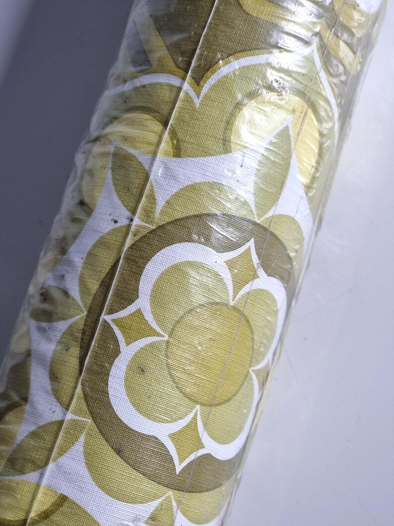 May include: A roll of wallpaper wrapped in clear plastic featuring a repeating geometric floral pattern in shades of olive green, mustard yellow, and white. The surface has a subtle textured finish with clean lines forming circular flower shapes.