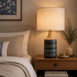 May include: A table lamp with a cylindrical ceramic base featuring a textured, multi-colored glaze in shades of blue, green, and brown. The lamp is topped with a large, off-white, drum-shaped fabric shade that emits a warm, soft glow.
