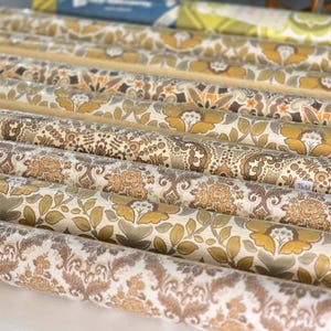 May include: A collection of multiple rolls of wallpaper arranged in parallel rows. Each roll features distinct patterns in shades of mustard yellow, brown, beige, and cream, including floral motifs, damask designs, and intricate botanical illustrations.