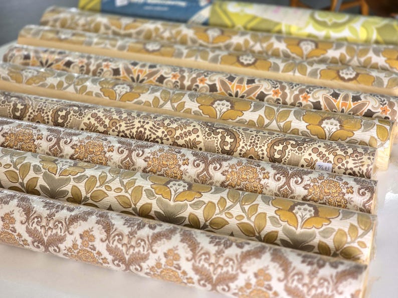 May include: A collection of multiple rolls of wallpaper arranged in parallel rows. Each roll features distinct patterns in shades of mustard yellow, brown, beige, and cream, including floral motifs, damask designs, and intricate botanical illustrations.