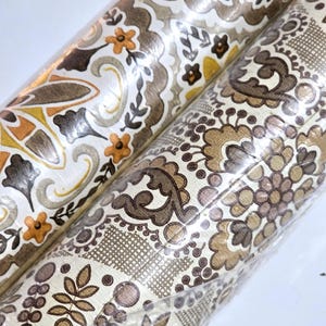 May include: Two rolls of wallpaper wrapped in clear plastic. The left roll features a repeating pattern of orange flowers and brown swirls on a cream background. The right roll displays a dense, intricate floral and geometric design in brown tones.