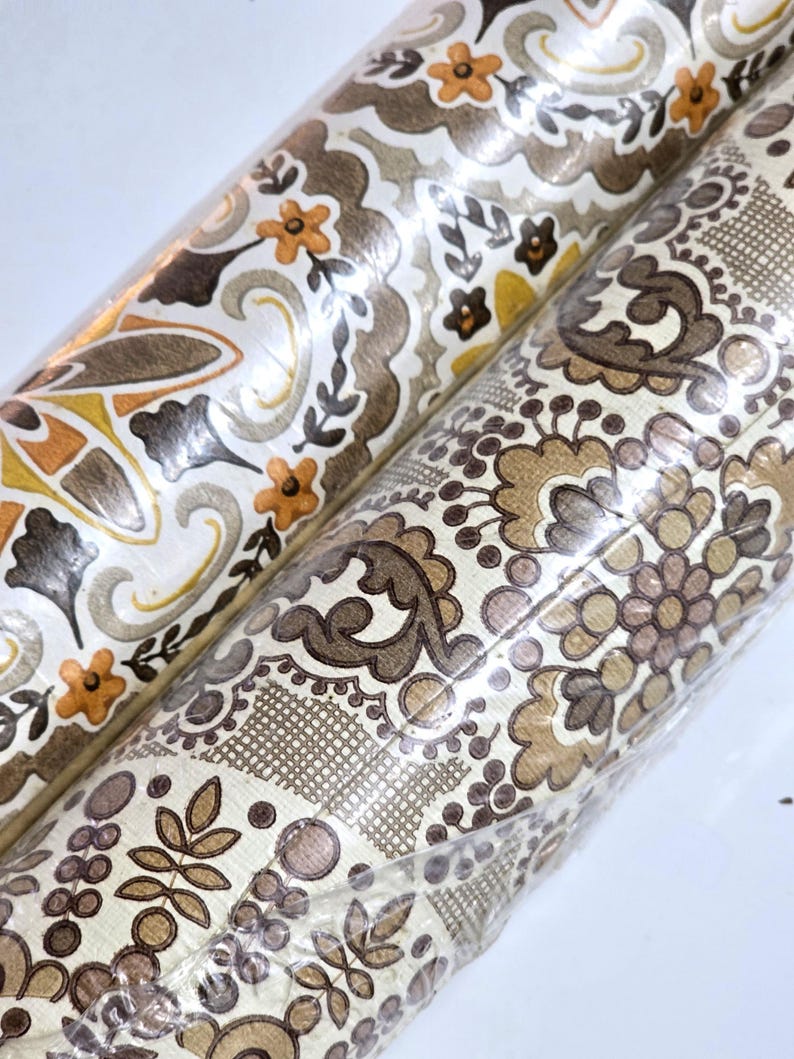 May include: Two rolls of wallpaper wrapped in clear plastic. The left roll features a repeating pattern of orange flowers and brown swirls on a cream background. The right roll displays a dense, intricate floral and geometric design in brown tones.