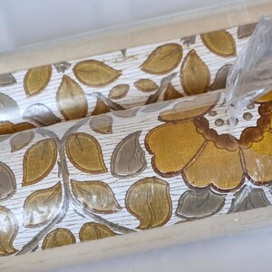 May include: A roll of wallpaper featuring a repeating pattern of large mustard yellow flowers and olive green leaves on a white background with a subtle wood grain texture. The roll is partially wrapped in clear plastic packaging.