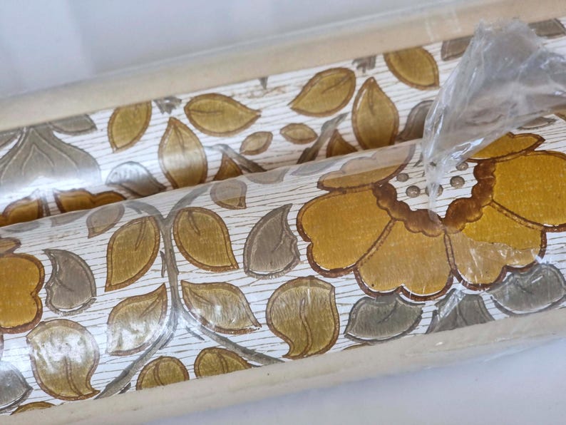 May include: A roll of wallpaper featuring a repeating pattern of large mustard yellow flowers and olive green leaves on a white background with a subtle wood grain texture. The roll is partially wrapped in clear plastic packaging.