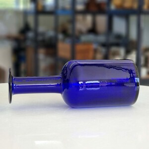 May include: A cobalt blue glass vase with a wide mouth and a narrow neck. The vase is lying on a white surface.