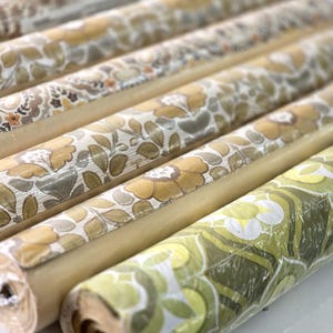 May include: A collection of rolled wallpaper featuring various floral and botanical patterns in shades of mustard yellow, olive green, brown, and cream. The rolls are arranged in parallel rows, showcasing repeating organic motifs and textures.