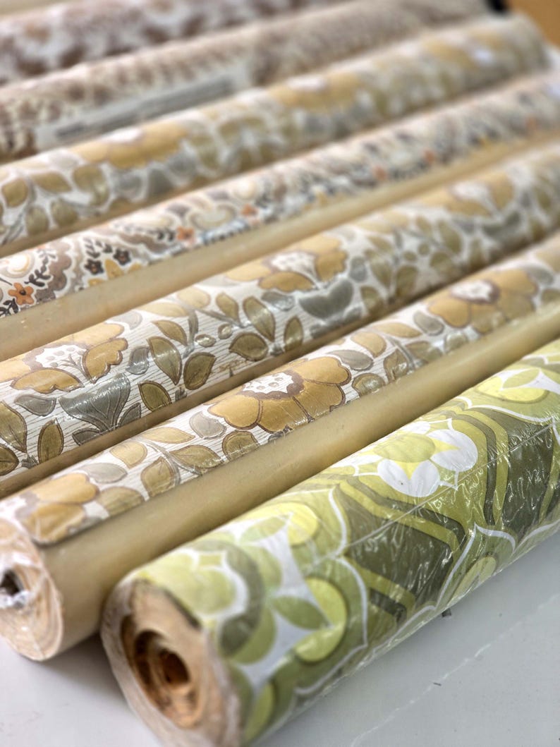 May include: A collection of rolled wallpaper featuring various floral and botanical patterns in shades of mustard yellow, olive green, brown, and cream. The rolls are arranged in parallel rows, showcasing repeating organic motifs and textures.