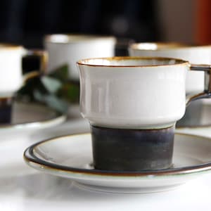 May include: Set of four white ceramic coffee cups with brown and black accents. The cups have a unique design with a wider base and a narrower top. Each cup is paired with a matching saucer.