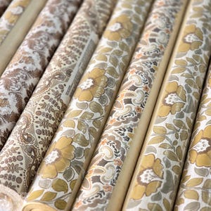 May include: Multiple rolls of wallpaper arranged in a row, each featuring distinct patterns in shades of mustard yellow, brown, beige, and white. The designs include floral motifs, intricate paisley swirls, and geometric dot patterns on paper.