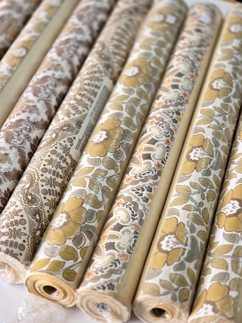 May include: Multiple rolls of wallpaper arranged in a row, each featuring distinct patterns in shades of mustard yellow, brown, beige, and white. The designs include floral motifs, intricate paisley swirls, and geometric dot patterns on paper.