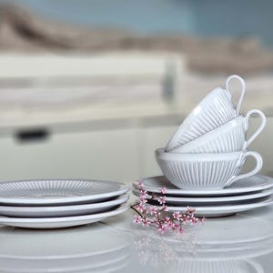 May include: A set of four white ceramic teacups stacked on top of each other. The cups have a ribbed design and are sitting on a white saucer. A small bouquet of pink flowers is in front of the cups.