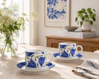 May include: Two white ceramic teacups and matching saucers featuring a blue floral pattern with green leaves. Each cup has a rounded handle and a wide rim. The saucers have a solid blue border, and the cups are decorated with blue blossoms.