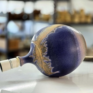 May include: A ceramic lamp base with a purple and gold glaze. The base is shaped like a sphere with a small neck and a white socket.