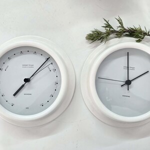 May include: Two white, round clocks with black hands and minimalist designs. One clock has a gauge with markings and the text "Siemens" and "MB". The other clock has only hour and minute hands. A sprig of greenery is in the background.