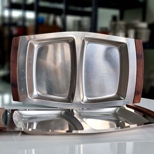May include: A stainless steel serving tray with two divided sections and wooden handles. The tray is made of polished metal and has a modern design.