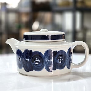 May include: A white ceramic teapot with a lid, featuring a dark blue floral pattern. The lid and rim have a dark blue band. The teapot has a curved spout and a rounded handle. A classic design for serving tea.