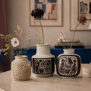May include: Three ceramic vases with relief carvings of figures. The left vase is light beige, the center vase is white with a dark brown base, and the right vase is dark brown with a white relief. Each holds delicate dried flowers and stems.