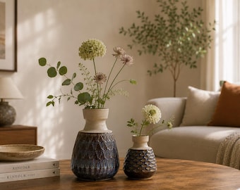 May include: Two ceramic vases with a two-tone design featuring a smooth cream-colored neck and a textured, dark blue and brown base with a diamond-shaped pattern. The vases are displayed with green stems and white flowers in each vessel.