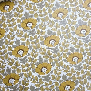 May include: A roll of wallpaper featuring a repeating floral pattern with mustard yellow flowers and grey-green leaves on a white textured background. The design consists of interconnected vines and foliage surrounding the stylized blossoms.