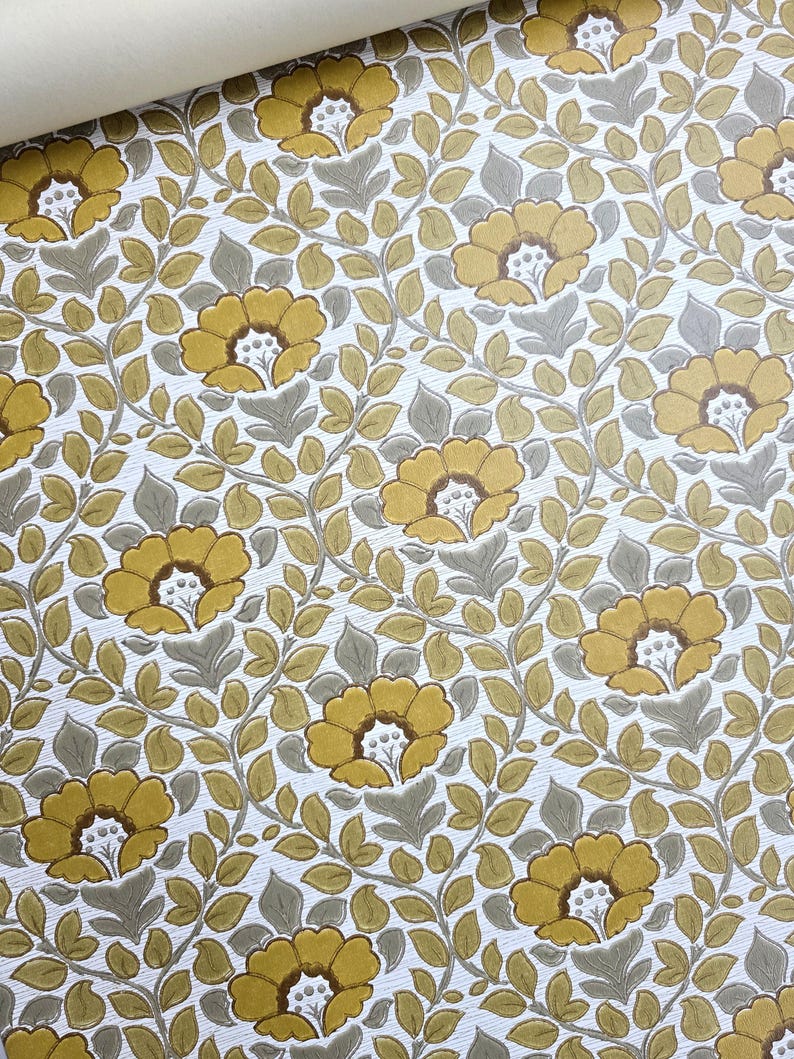 May include: A roll of wallpaper featuring a repeating floral pattern with mustard yellow flowers and grey-green leaves on a white textured background. The design consists of interconnected vines and foliage surrounding the stylized blossoms.