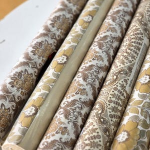 May include: Several rolls of wallpaper featuring a repeating floral and paisley pattern in shades of mustard yellow, brown, and cream. The rolls are arranged side-by-side, showcasing the intricate botanical motifs and textured surface details.