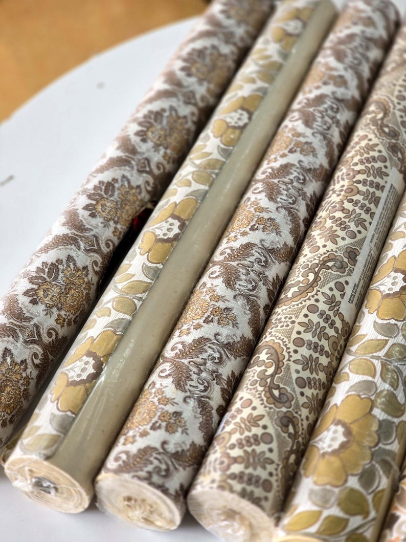 May include: Several rolls of wallpaper featuring a repeating floral and paisley pattern in shades of mustard yellow, brown, and cream. The rolls are arranged side-by-side, showcasing the intricate botanical motifs and textured surface details.