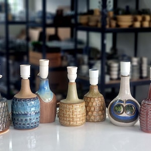 May include: A selection of seven ceramic table lamps with diverse designs and colours. The lamps have white lamp holders and are displayed on a white surface. The lamps feature patterns, textures, and a range of colours, including blue, brown, and beige.