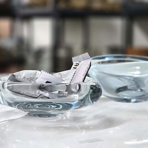 May include: Two clear glass bowls, one with a crumpled white and brown wrapper inside. The bowls are on a white surface.