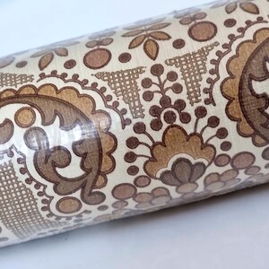 May include: A roll of wallpaper featuring a repeating pattern of brown and tan paisley-inspired floral motifs, circular shapes, and cross-hatched triangular elements on a cream-colored background, all encased in a clear plastic protective film.