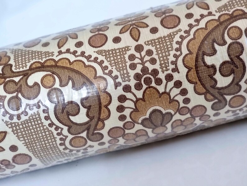 May include: A roll of wallpaper featuring a repeating pattern of brown and tan paisley-inspired floral motifs, circular shapes, and cross-hatched triangular elements on a cream-colored background, all encased in a clear plastic protective film.