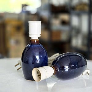May include: Two ceramic table lamps with a glossy, dark blue glaze. Each lamp has a white socket and a brown band near the base of the socket. One lamp is upright, while the other is lying on its side. The lamps are on a white surface.