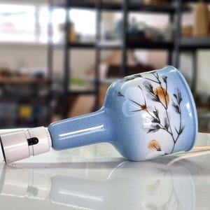 May include: A light blue ceramic lamp with a floral design. The lamp has a white bulb socket and a white cord. The floral design features orange and white flowers with black stems and leaves.