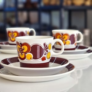 May include: Three brown and yellow floral teacups and saucers. The cups have a brown rim and a white interior. The saucers have a brown rim and a white centre.