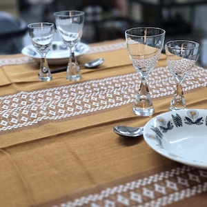 May include: A table setting with four clear glass wine glasses, a white plate with a blue and white floral design, and a silver spoon on a brown tablecloth with a white geometric pattern.
