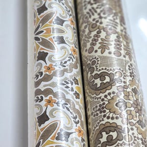 May include: Two rolls of wallpaper featuring intricate patterns. The left roll displays a repeating floral motif in shades of mustard yellow, brown, and cream. The right roll features a dense, detailed paisley and lace-like design in brown tones.