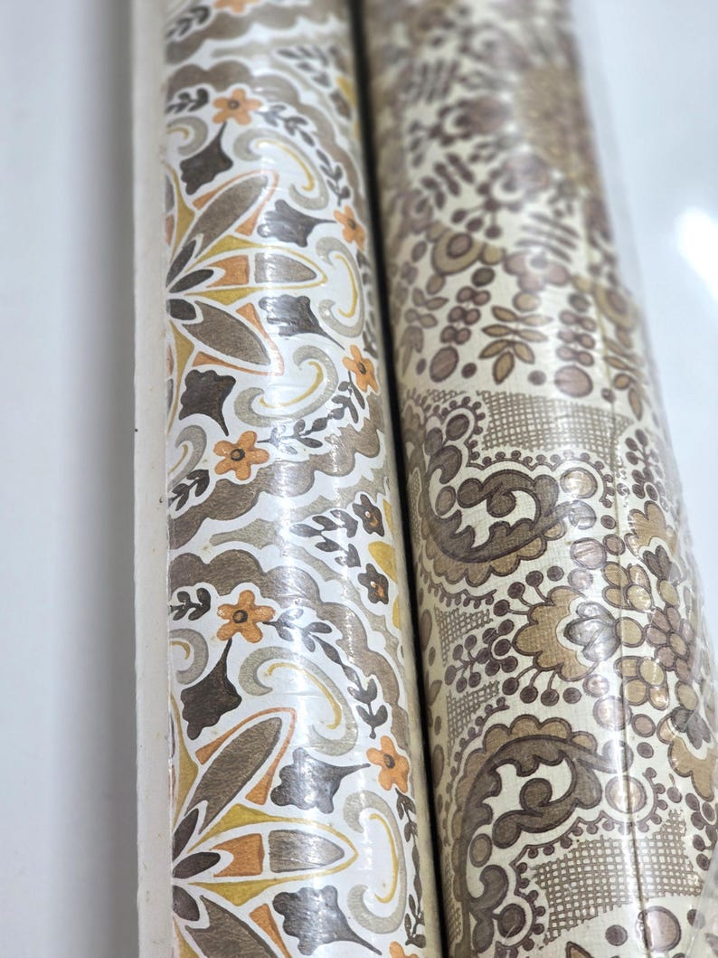 May include: Two rolls of wallpaper featuring intricate patterns. The left roll displays a repeating floral motif in shades of mustard yellow, brown, and cream. The right roll features a dense, detailed paisley and lace-like design in brown tones.