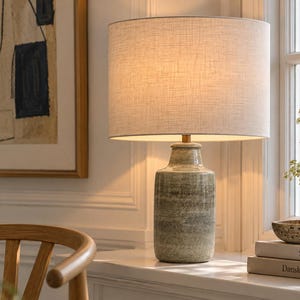 May include: A table lamp with a cylindrical, mottled green ceramic base and a light-coloured, textured fabric drum shade. The lamp emits a warm glow and sits next to two stacked books labelled SØHOLM and Dansk keramik near a window.