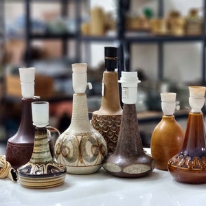 Puede incluir: Siete lámparas de mesa de cerámica con diferentes diseños y colores. Las lámparas son todas de un estilo similar, con una base ancha y un cuello estrecho. Las lámparas son todas en varios tonos de marrón, verde y blanco.