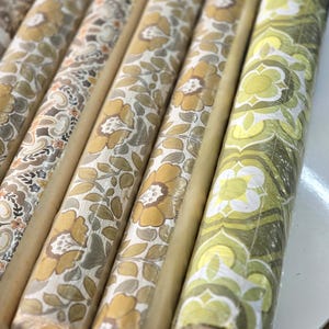 May include: A collection of cylindrical wallpaper rolls arranged side by side. The rolls feature various patterns including large yellow and beige floral motifs, intricate paisley designs, and a repeating geometric green and white flower print.