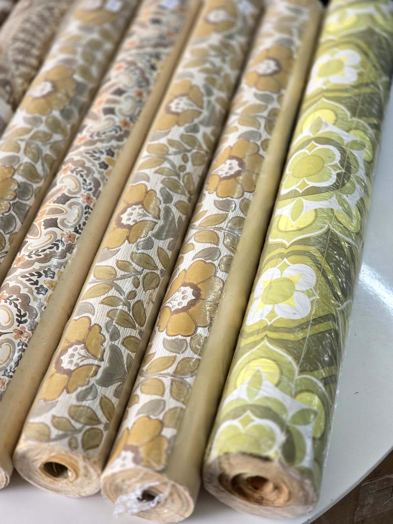 May include: A collection of cylindrical wallpaper rolls arranged side by side. The rolls feature various patterns including large yellow and beige floral motifs, intricate paisley designs, and a repeating geometric green and white flower print.