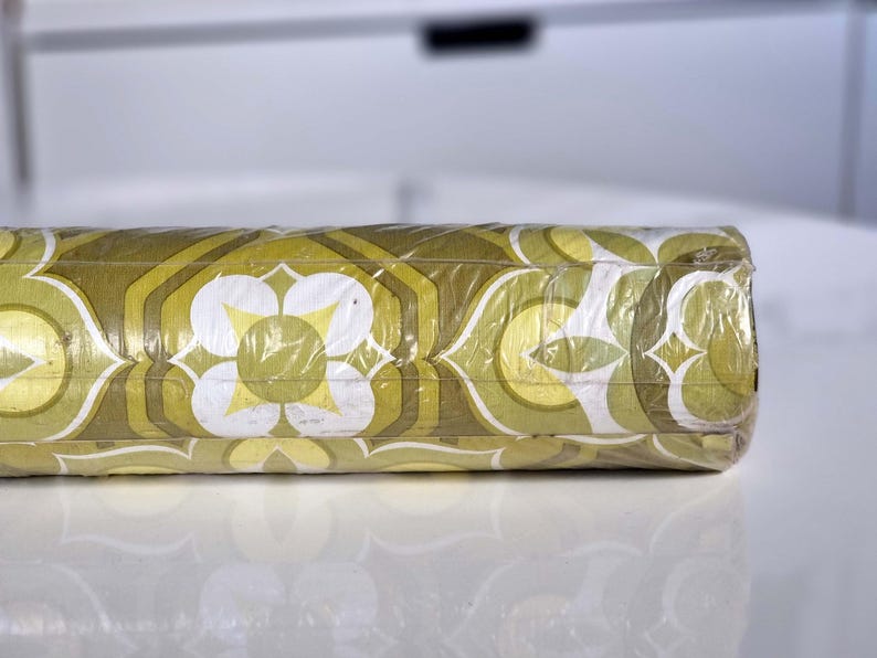 May include: A roll of wallpaper wrapped in clear plastic film, featuring a repeating geometric pattern in shades of olive green, lime green, and white. The design consists of stylized floral shapes and interlocking hexagonal outlines.