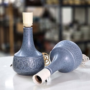 Op de afbeelding: Twee blauwe keramische lampvoeten met een gespikkelde afwerking en een herhalend reliëf geometrisch patroon rond de onderkant. Elke lampvoet heeft een hoge, slanke hals met een witte plastic fitting en een bijbehorend wit snoer.