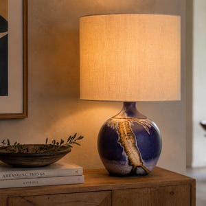 Op de afbeelding: Een tafellamp met een cilindrische beige stoffen kap en een bolvormige keramische voet met een diepblauw glazuur en een gestructureerd, verticaal bruin en crème craquelépatroon in het midden van de ronde behuizing.