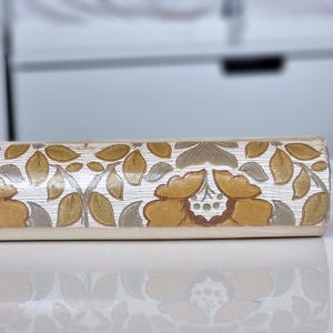May include: A cylindrical rolling pin featuring a repeating floral pattern with mustard yellow, brown, and muted green leaves and petals against a white background. The surface has a textured, wood-grain appearance with carved details.